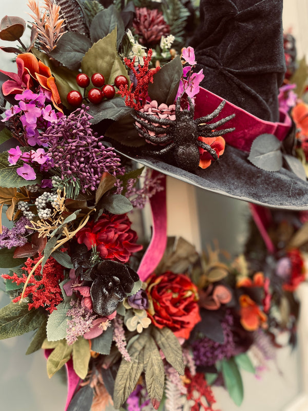 Halloween-themed wreath with dark green leaves, red and orange flowers, and a black witch hat.
