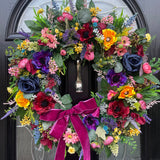 An artificial luxury faux floral wreath with bright jewel tones including deep purple, navy blue, burgundy, pink and bright yellow. Finished off with a velvet bow.