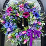 A large artificial door wreath with a vibrant arrangement of purple, pink, and blue anemones and hydrangea, accented with a purple ribbon.